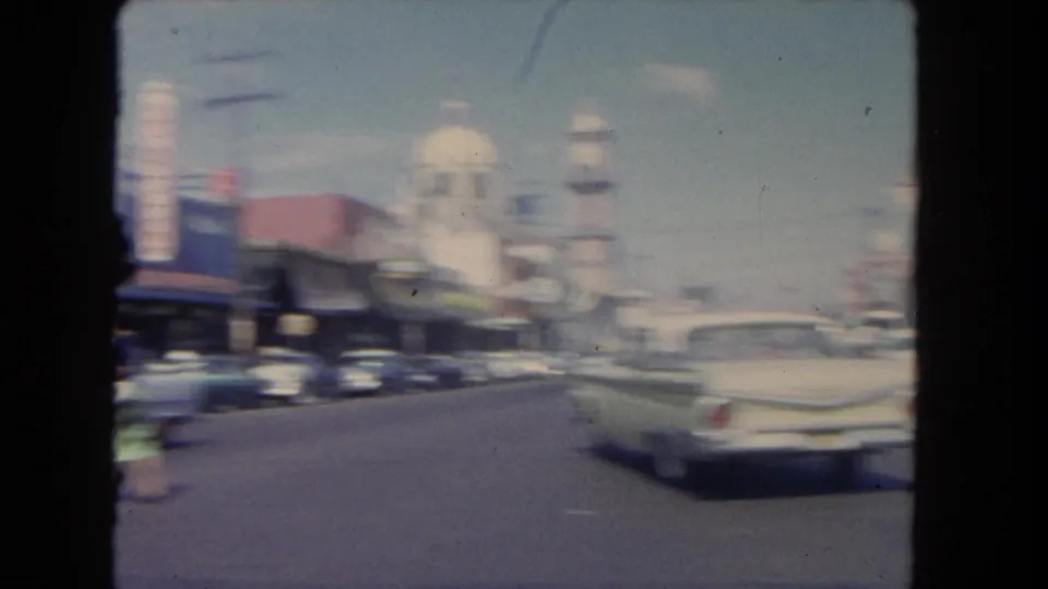 A moving 8mm view of a Los Angeles city street with parked cars, storefronts, and a clock-tower-like structure in the distance.