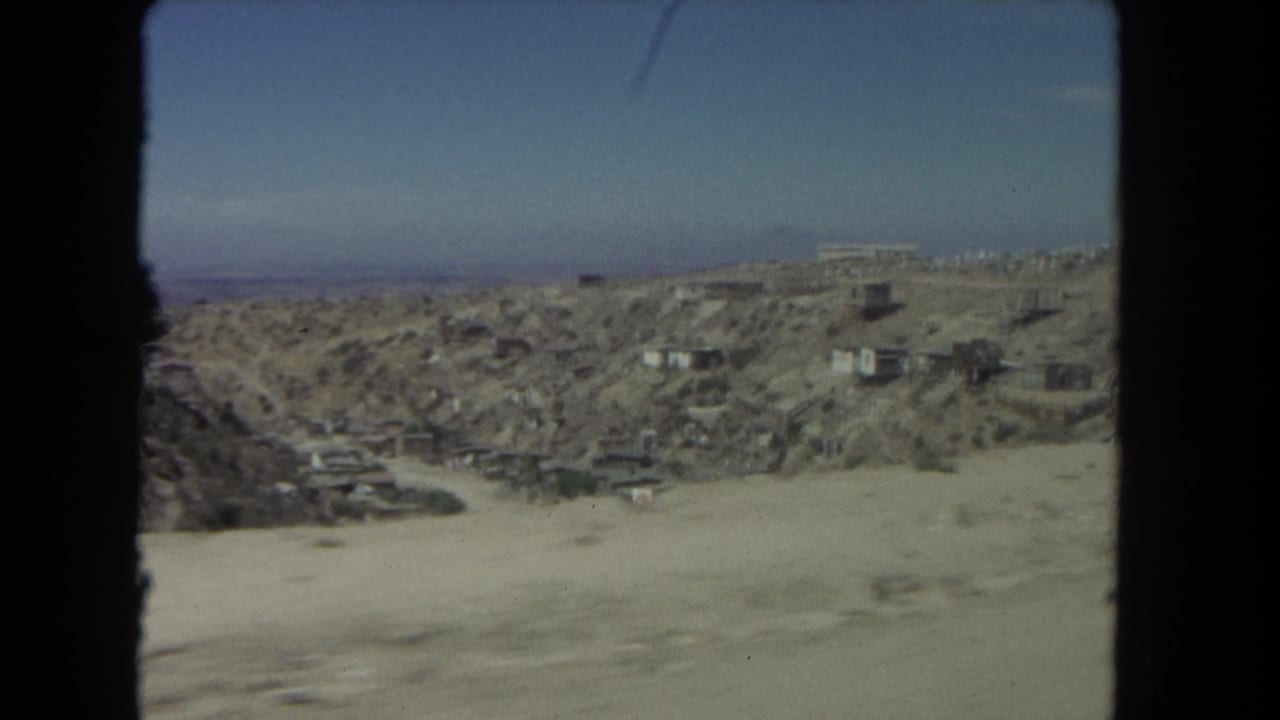1962 Los Angeles roadside view showing an arid hillside, small structures, and distant city edge through 8mm film blur.