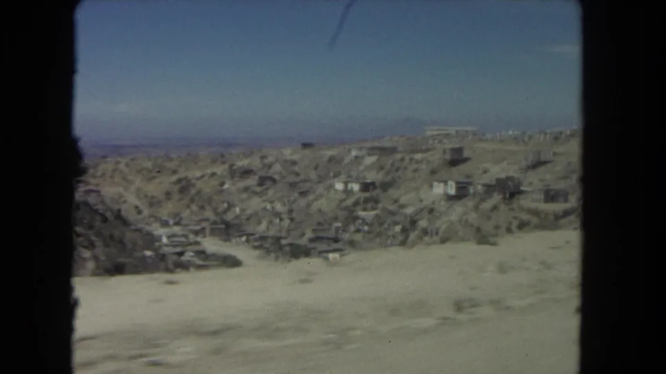 1962 Los Angeles roadside view showing an arid hillside, small structures, and distant city edge through 8mm film blur.