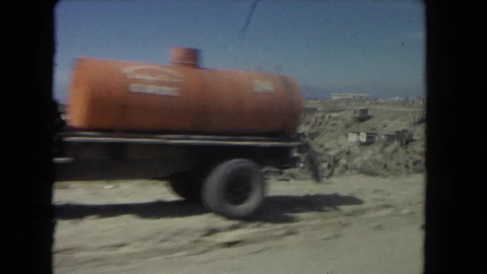 1962 Los Angeles driving footage frame with an orange tanker truck passing across a dry hillside roadside scene.