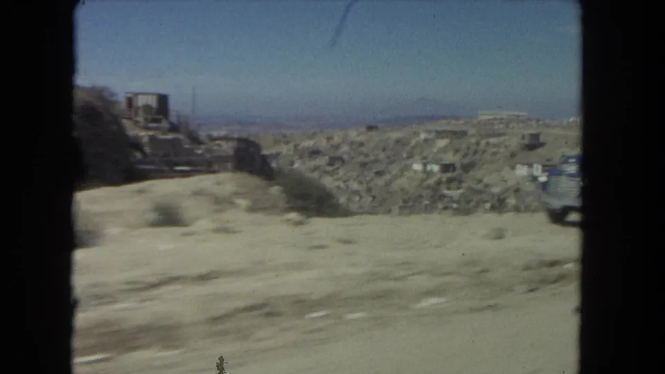 1962 Los Angeles home-movie frame showing a dusty roadside, hillside structures, and a partial vehicle at the right edge.