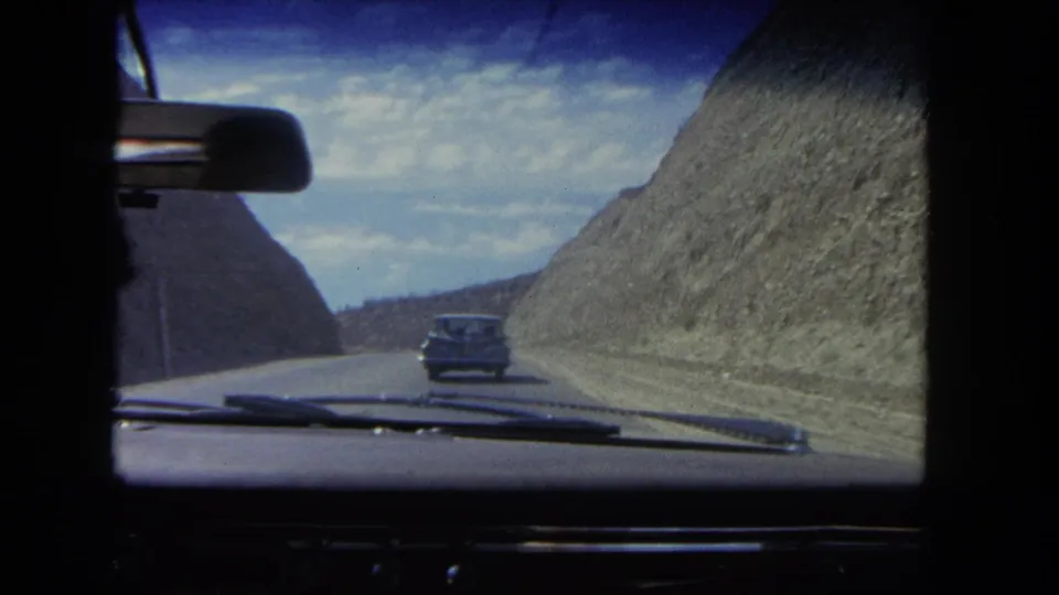 1962 8mm windshield view near Los Angeles showing a dark sedan ahead on a paved road cut through rocky hills.