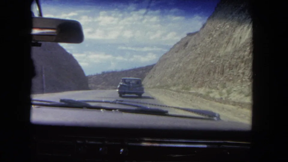 Vintage 8mm road footage from inside a car, with a sedan ahead and dry rocky slopes beside the roadway.