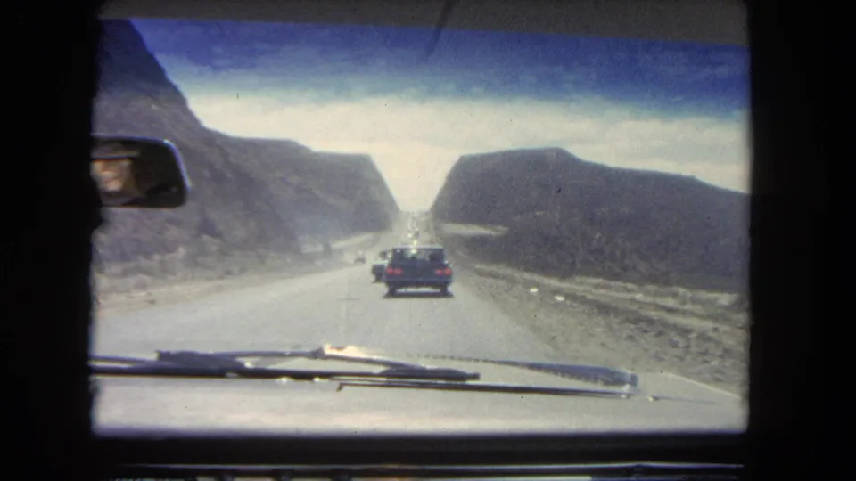 1962 Los Angeles-area driving view through a windshield, showing a bright road through a rocky cut and a car ahead.