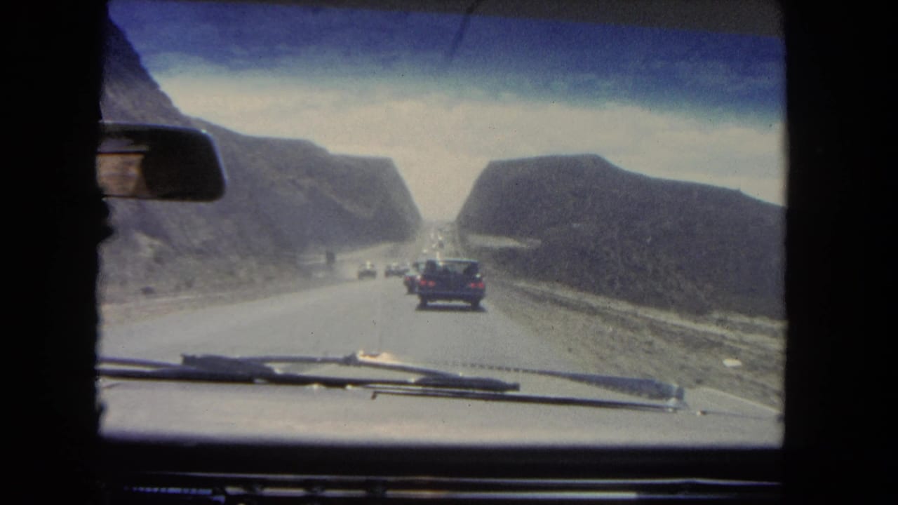 Vehicle windshield view of a car ahead on a road between hills.
