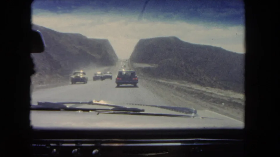 Windshield-shot 1962 California road scene with cars ahead and oncoming traffic in a dry mountain pass.
