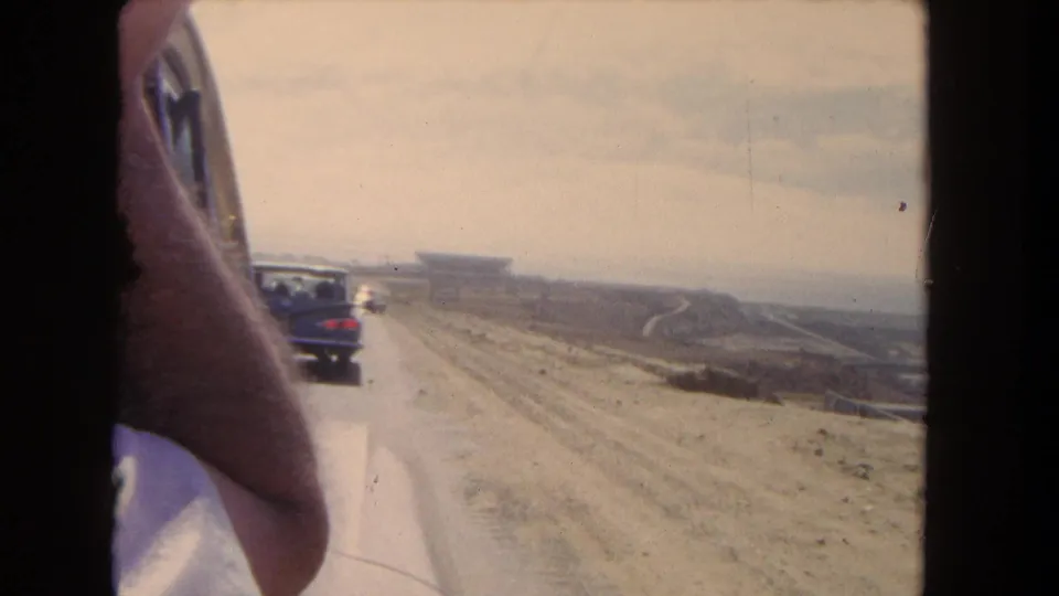 1962 Los Angeles-area 8mm frame from inside a moving vehicle, showing a forearm at the window, cars ahead, and an arid roadside landscape.