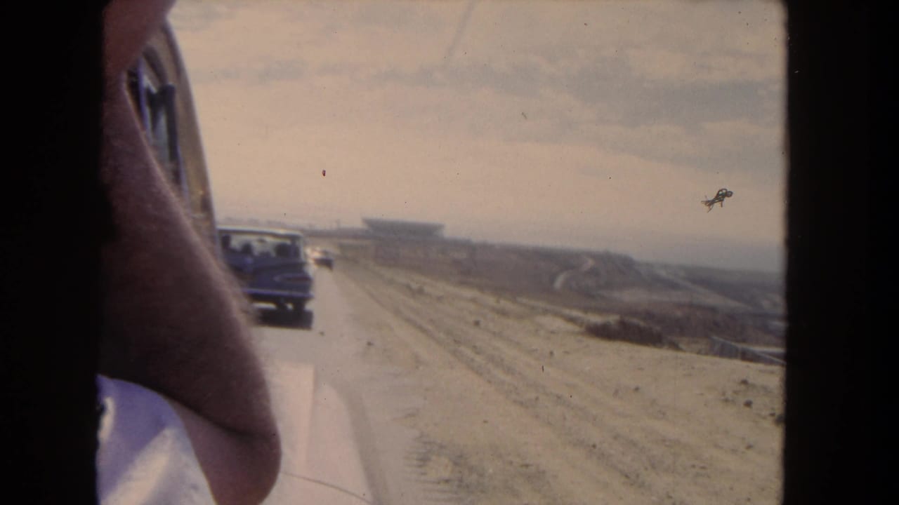 Moving vehicle view with an arm at the window, cars on the road, and a hazy structure in the distance.