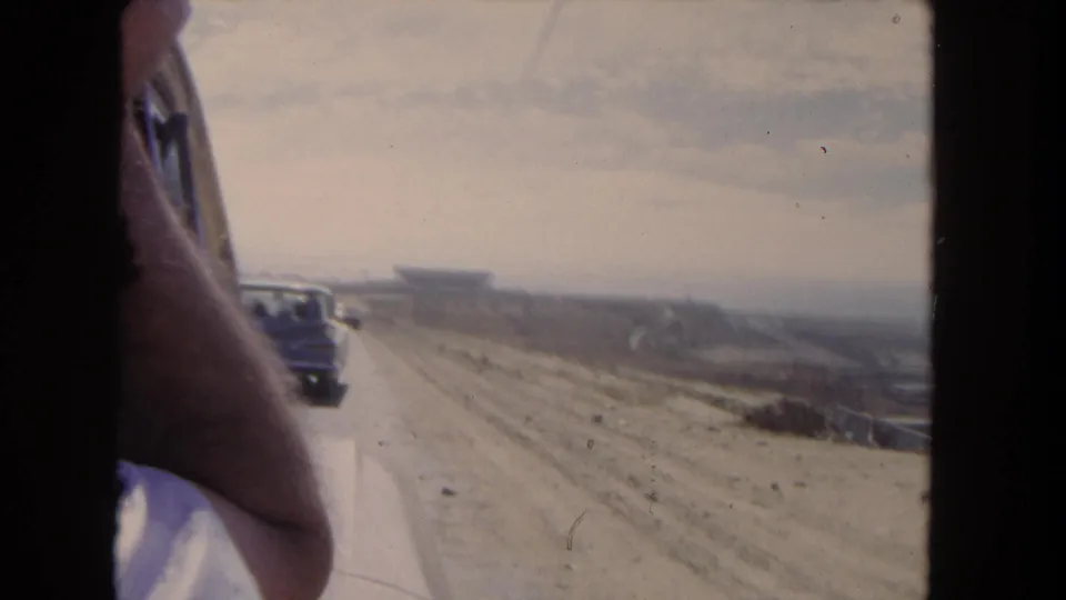Final 1962 Los Angeles-area frame from the moving vehicle, with the passenger arm, road traffic, arid shoulder, and distant structure visible.