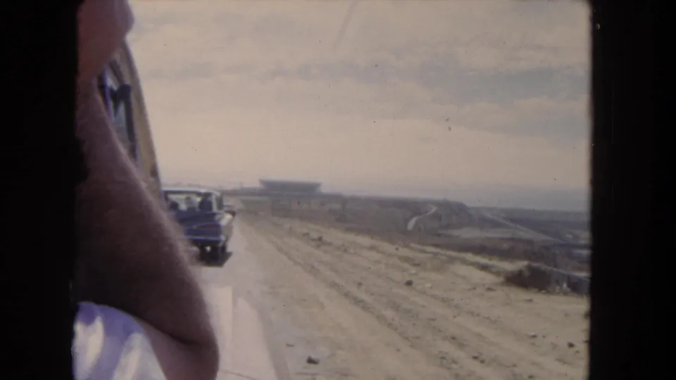 1962 Los Angeles home-movie view from inside a vehicle, with a passenger at left and a period car ahead on an unpaved road.