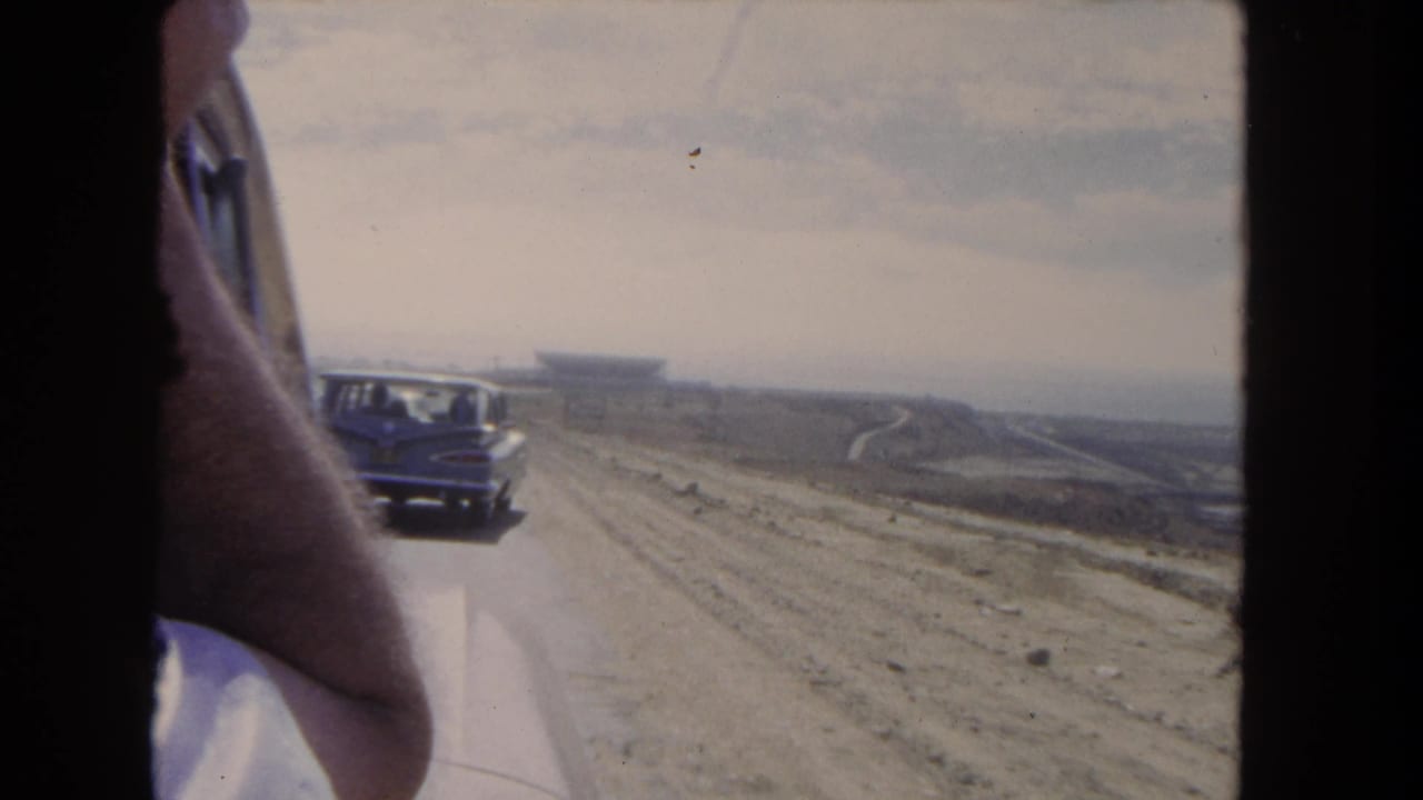 Car ahead on a rough dirt road with open land and a distant structure.