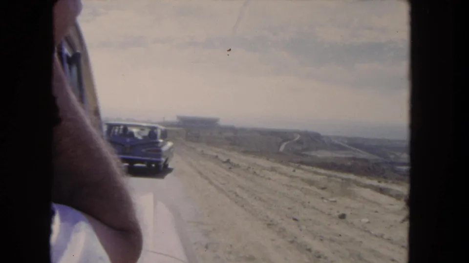 Home-movie frame from a moving car in 1962 Los Angeles, showing a period automobile on an unpaved road across open terrain.