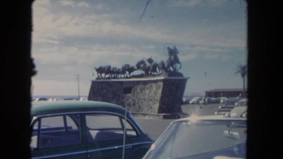 1962 Los Angeles roadside monument scene with parked period cars and a bronze-looking cowboy-and-cattle sculpture on a stone base.