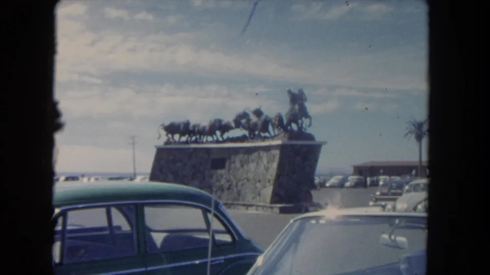 Passing 1962 Los Angeles view of a stone monument with cowboy-and-cattle sculpture, parked cars, and a palm at the edge of frame.