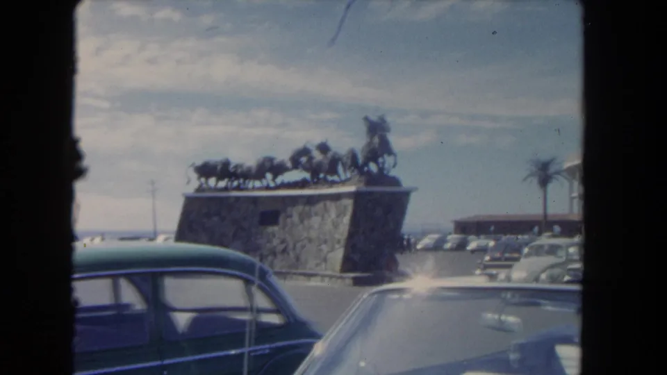 1962 Los Angeles home-movie frame showing parked mid-century cars beside a stone monument with a cowboy-and-horse sculpture.