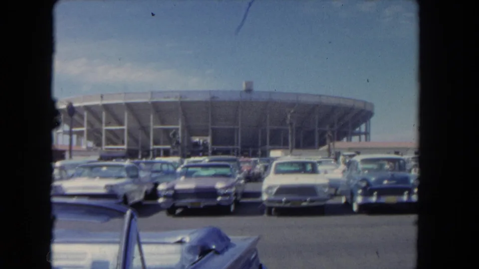 1962 Los Angeles home-movie frame with several parked sedans and a blue car before a curved stadium or arena exterior.