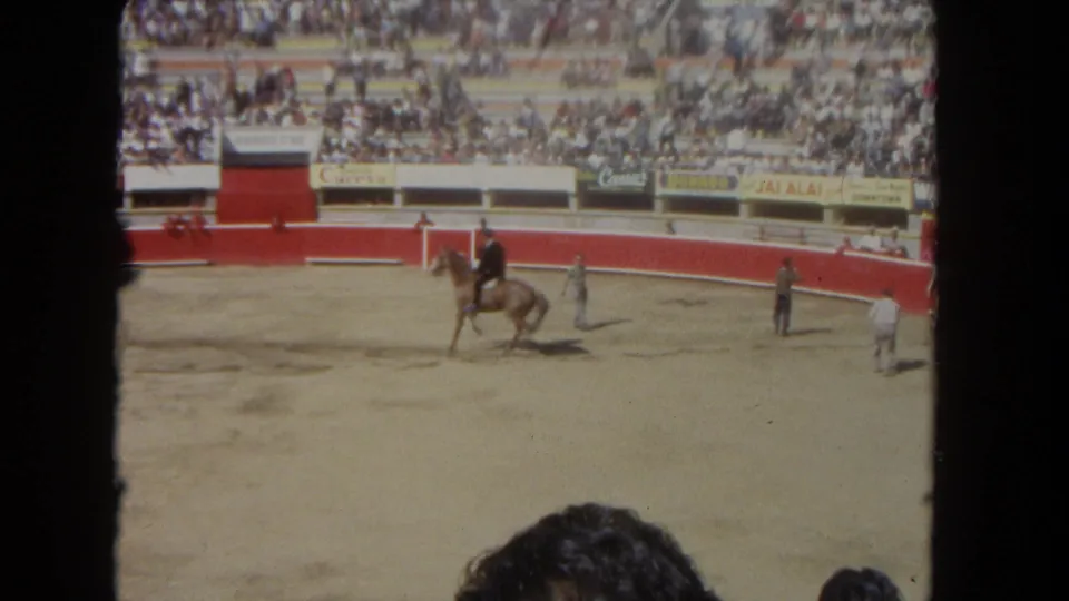 Wide 8mm view of a rider and horse crossing a rodeo arena with staff on the floor and crowded stands beyond.