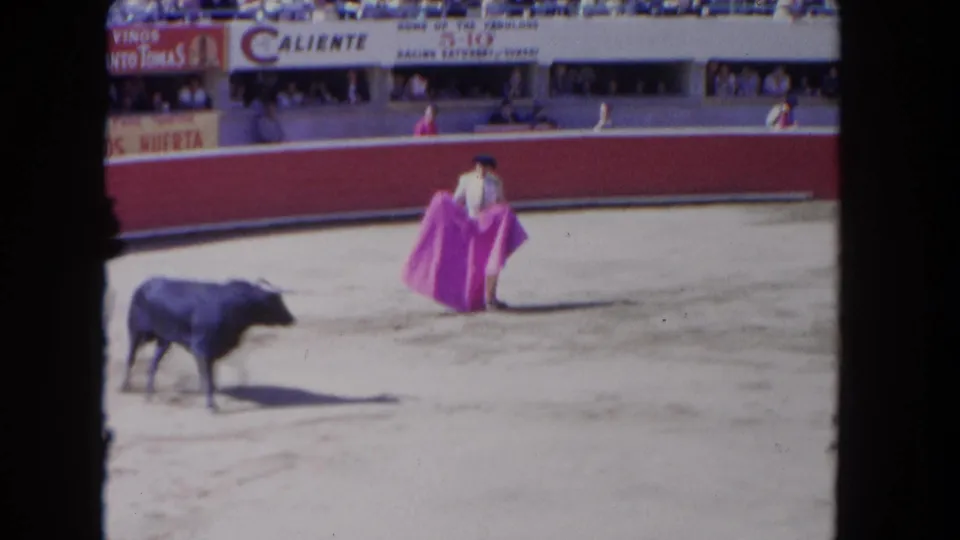 1962 Los Angeles 8mm frame showing a matador with a pink cape facing a black bull before spectators and arena signage.