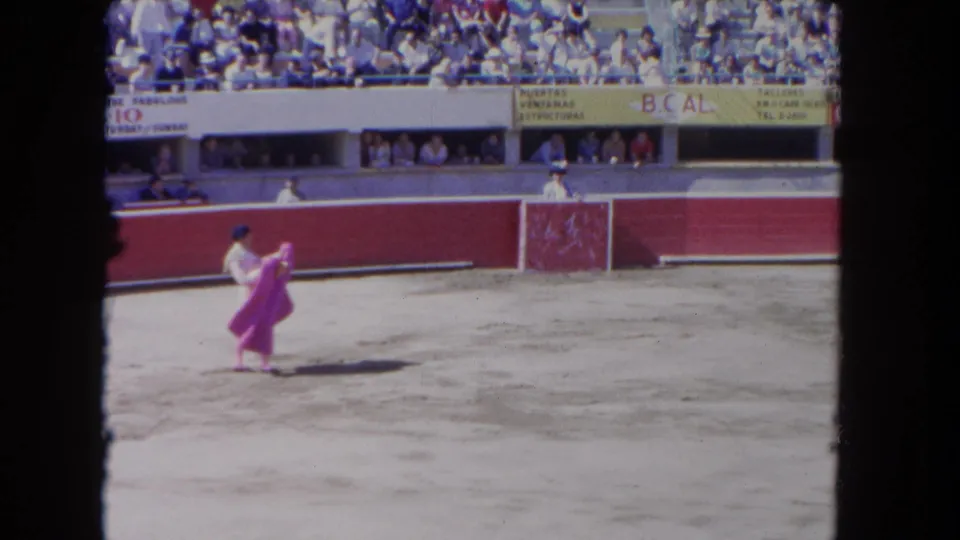 The 8mm frame shows the matador repositioning the pink cape while spectators watch from the stands behind the red barrier.