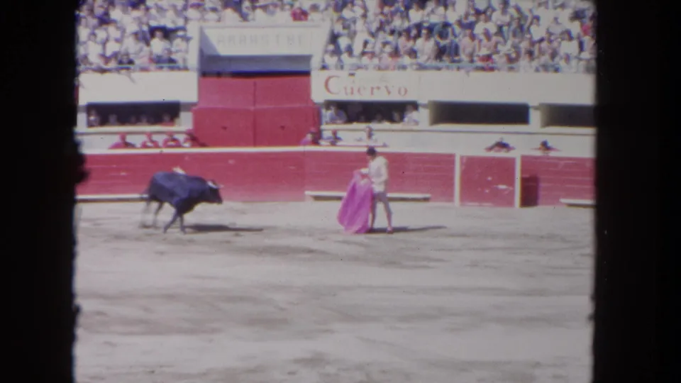 1962 Los Angeles home-movie frame showing a matador holding a pink cape as a dark bull stands nearby in a red-walled arena.