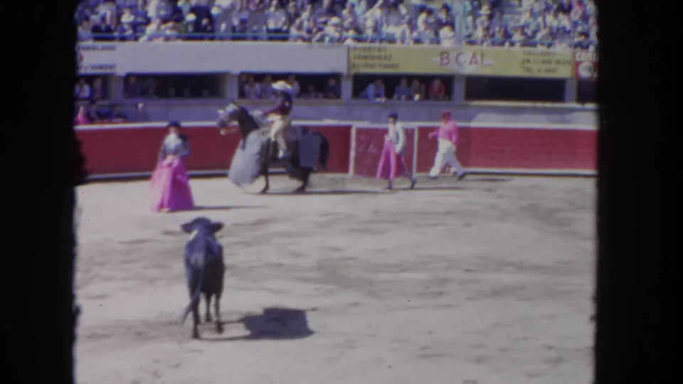 1962 Los Angeles bullring frame with a dark bull in the foreground, a mounted rider, assistants, and spectators behind the red wall.