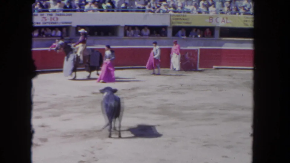 1962 Los Angeles 8mm frame showing a dark bull in a sandy arena as matadors and a mounted rider stand near red barriers.