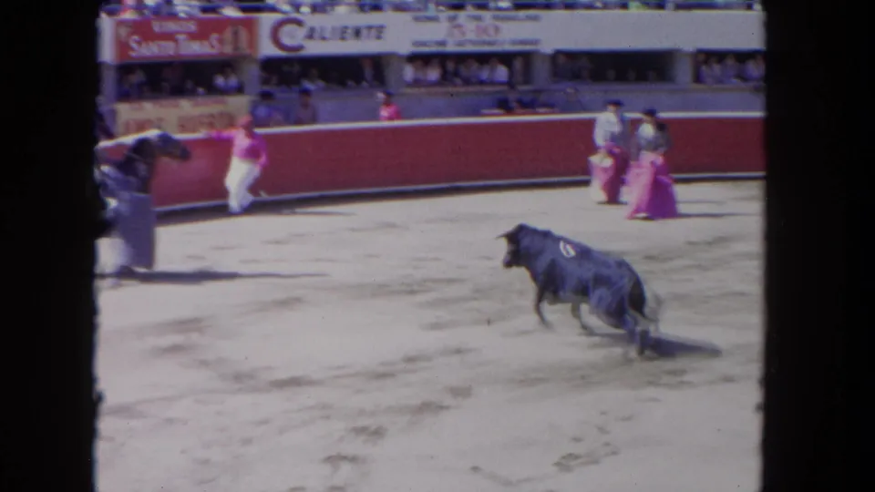 1962 Los Angeles 8mm frame of a dark bull running across the arena while caped performers wait near the red barrier.