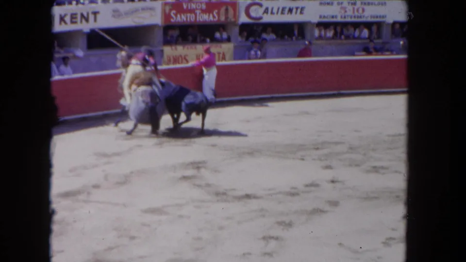 1962 Los Angeles bullfight scene with a mounted rider close to the bull and a matador signaling beside the red arena barrier.