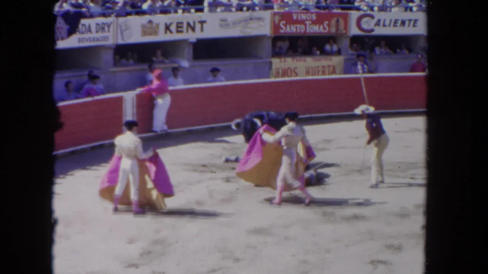 1962 Los Angeles 8mm frame with matadors in white suits and bright capes reacting as the bull lowers its head near the arena floor.