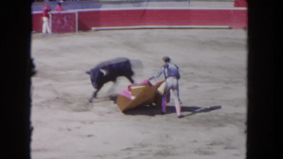 1962 Los Angeles 8mm frame showing a matador using a pink and yellow cape near a black bull in a sunlit arena.