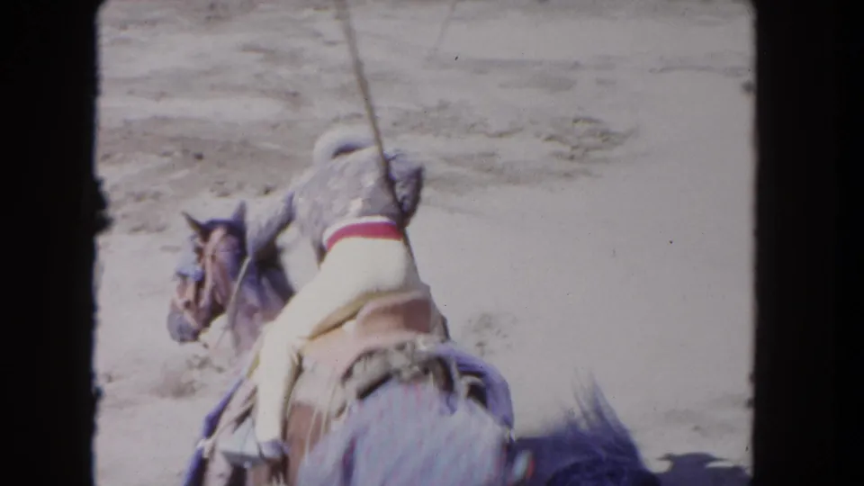 1962 Los Angeles 8mm frame of a mounted cowboy in pale trousers and a red sash riding through a dusty arena with a lance.