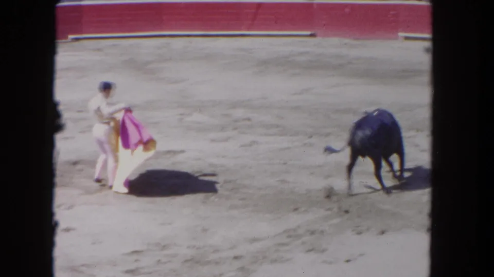 1962 Los Angeles home-movie frame showing a bullfighter with a pink cape facing a dark bull inside a red-walled arena.