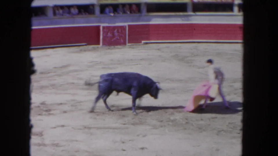 1960s 8mm frame showing a bull approaching a matador-style performer with a pink cape inside a bullring.