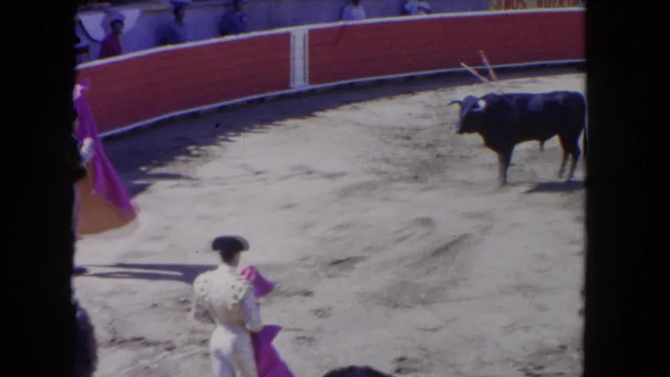 8mm film circa 1962 — close view of matador displaying the muleta cape; dark bull bearing banderillas visible at ring-right in afternoon sun, red barrier wall curves behind.