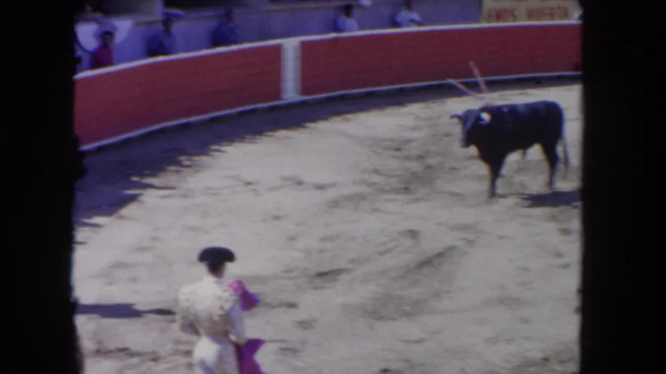 8mm home-movie frame, c.1962: matador in white embroidered suit faces black bull across sandy bullring; spectators behind red barrera, arena signage visible at top.