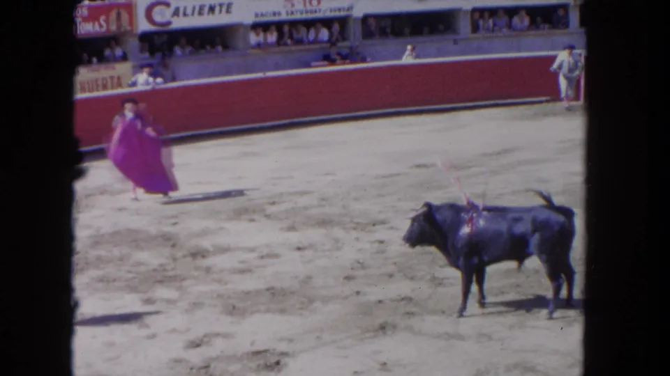 8mm home-movie frame, c.1962: matador works bull bearing what appear to be banderillas in sun-drenched arena; 'Caliente' and Spanish-language boards legible in crowded stands.