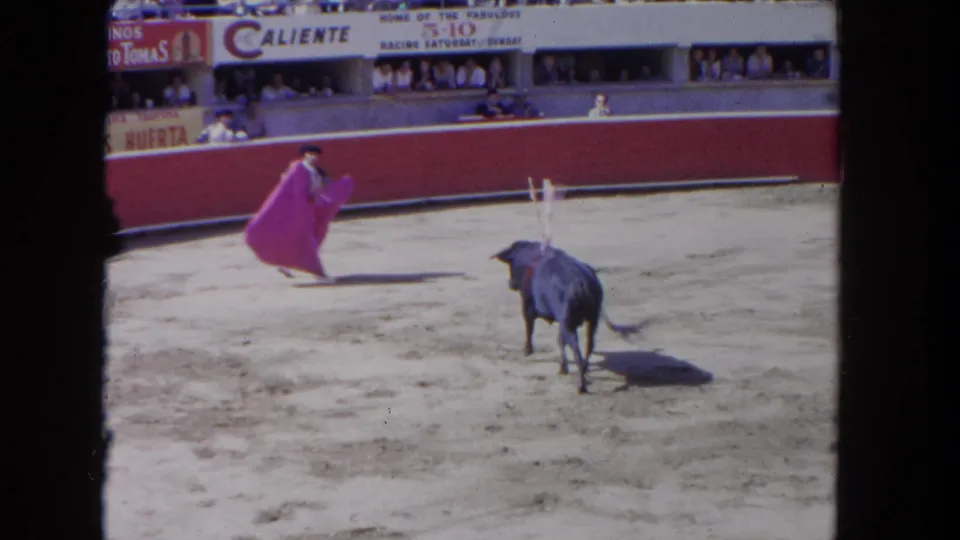 8mm home-movie frame, c.1962: close cape pass with charging bull; hard midday shadow on sand; 'Caliente' and 'Huerta' signage clearly legible above red barrera and crowd.