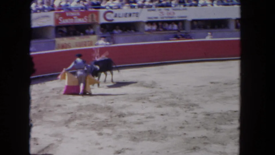 8mm home-movie frame, c.1962: matador at very close range with bull near the barrera; cape and possible muleta cloth in motion; 'Caliente' and Vinos Santo Tomás-style boards in background.