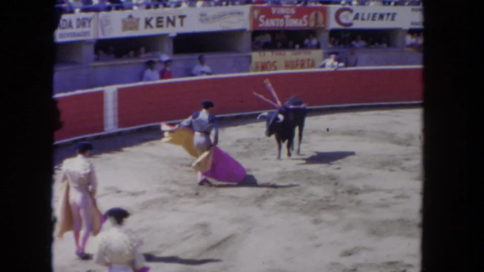 8mm home movie frame circa 1962 showing three matadors in cream traje de luces working a dark bull with banderillas; Kent cigarette and Caliente banners above packed grandstands.