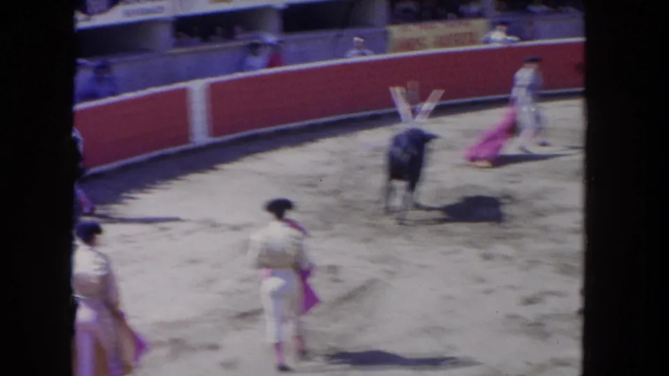 8mm action frame circa 1962 capturing a bull mid-charge toward a matador with a pink cape; banderillas remain planted in the bull's shoulders, multiple ring figures visible on the sand.