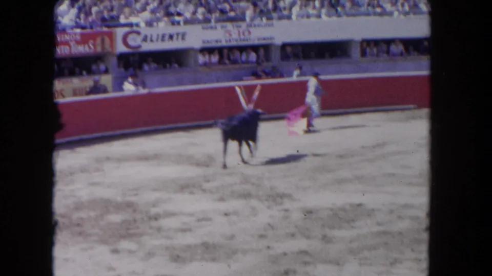 8mm spectator-shot frame circa 1962 of a matador and bull near the red barrera; Caliente and Vinos Santo Tomas banners prominent above densely packed grandstands in bright sunlight.