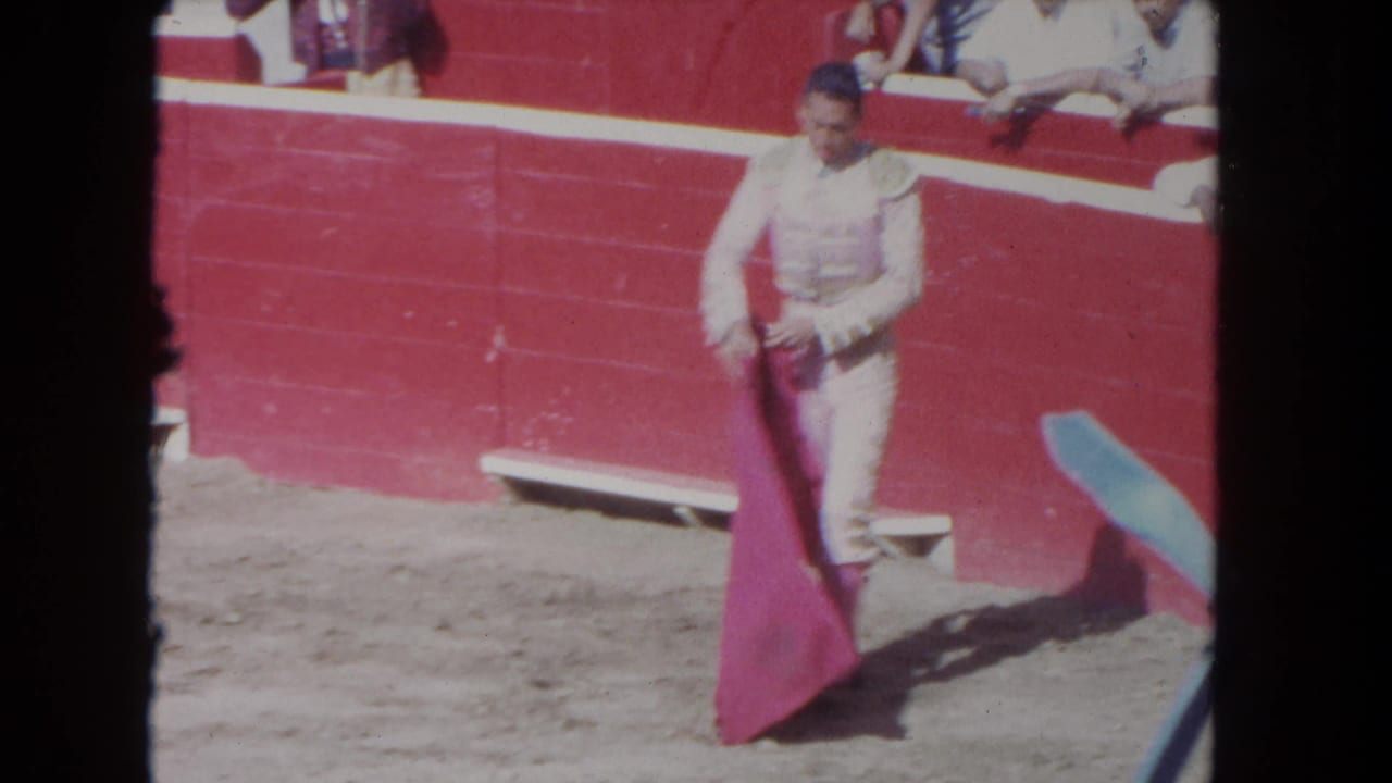 Full-body view of a matador with cape extended in the sand arena, circa 1962, red barrier wall and spectators in background.