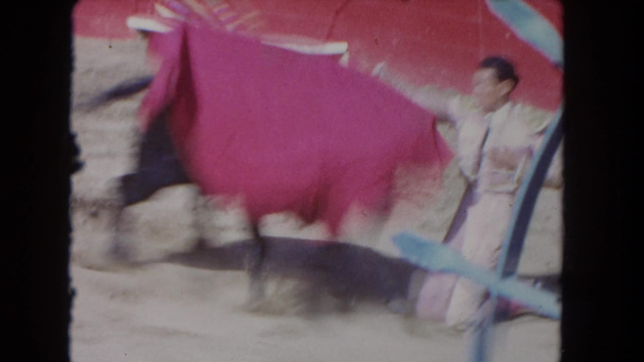8mm home-movie frame circa 1962 — matador mid-pass with magenta cape, sandy ring and red-painted arena wall behind, shot from behind a blue X-brace barrier.