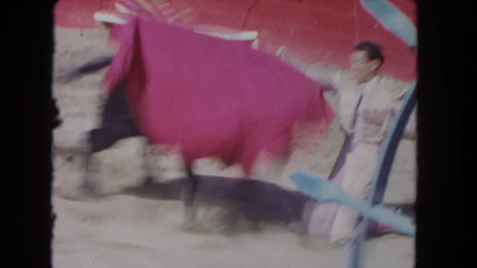 8mm home-movie frame circa 1962 — matador mid-pass with magenta cape, sandy ring and red-painted arena wall behind, shot from behind a blue X-brace barrier.