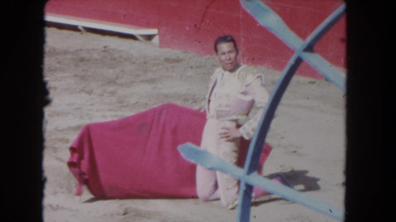 8mm frame circa 1962 — matador pausing between passes with cape at rest, red arena wall and white bench visible through blue X-frame barrier.