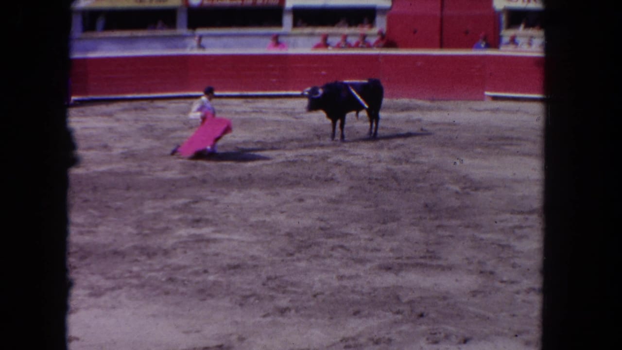 Wide shot of matador in traditional traje de luces extending red muleta toward dark bull; arena barrier with advertising signage and spectators visible, consistent with early 1960s 8mm color footage.