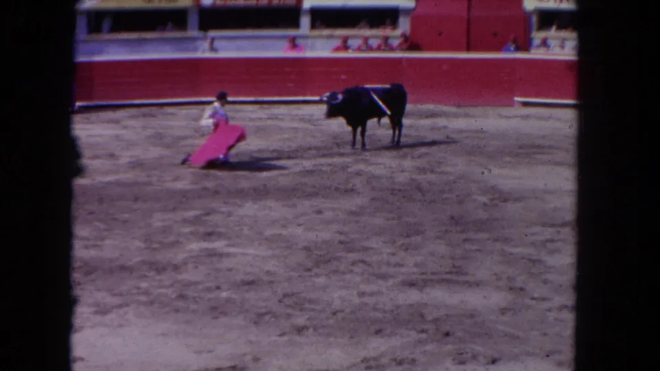 Wide shot of matador in traditional traje de luces extending red muleta toward dark bull; arena barrier with advertising signage and spectators visible, consistent with early 1960s 8mm color footage.