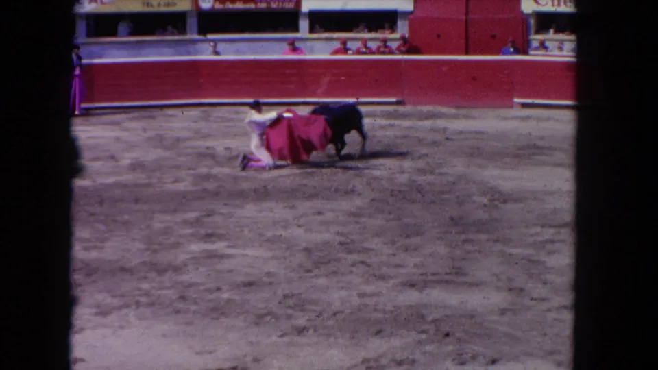 Matador kneeling with extended red muleta as dark bull charges; advertising boards on red arena barrier and a second pink-costumed figure nearby visible, early 1960s 8mm color footage.