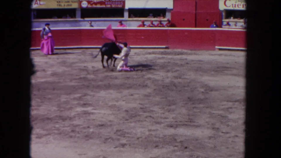 8mm film frame circa 1962 showing a matador in white standing close to a black bull in a bullfighting arena, pink-caped banderillero at left, partial advertising banners overhead.