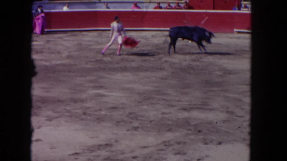8mm film frame circa 1962 capturing a matador sweeping a red cape as a black bull charges in a sunlit open arena, shot from the spectator stands.